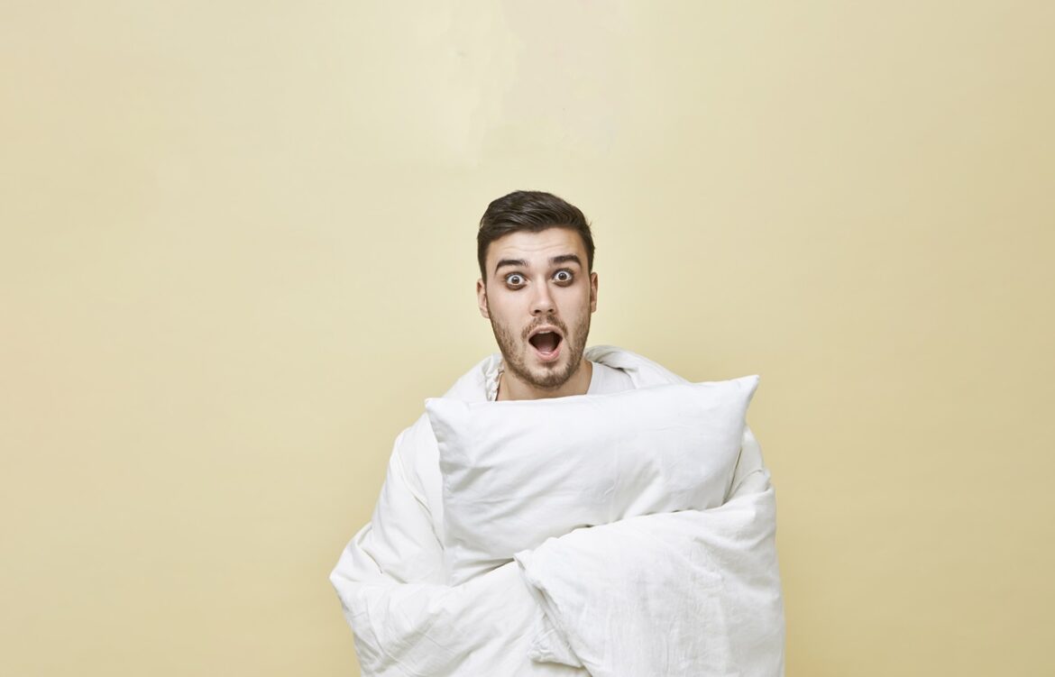 Isolated shot of terrified young bearded male rolling himself in white blanket and shouting with mouth wide opened, can’t fall asleep because of scary movie. Bedtime, sleep and insomnia concept